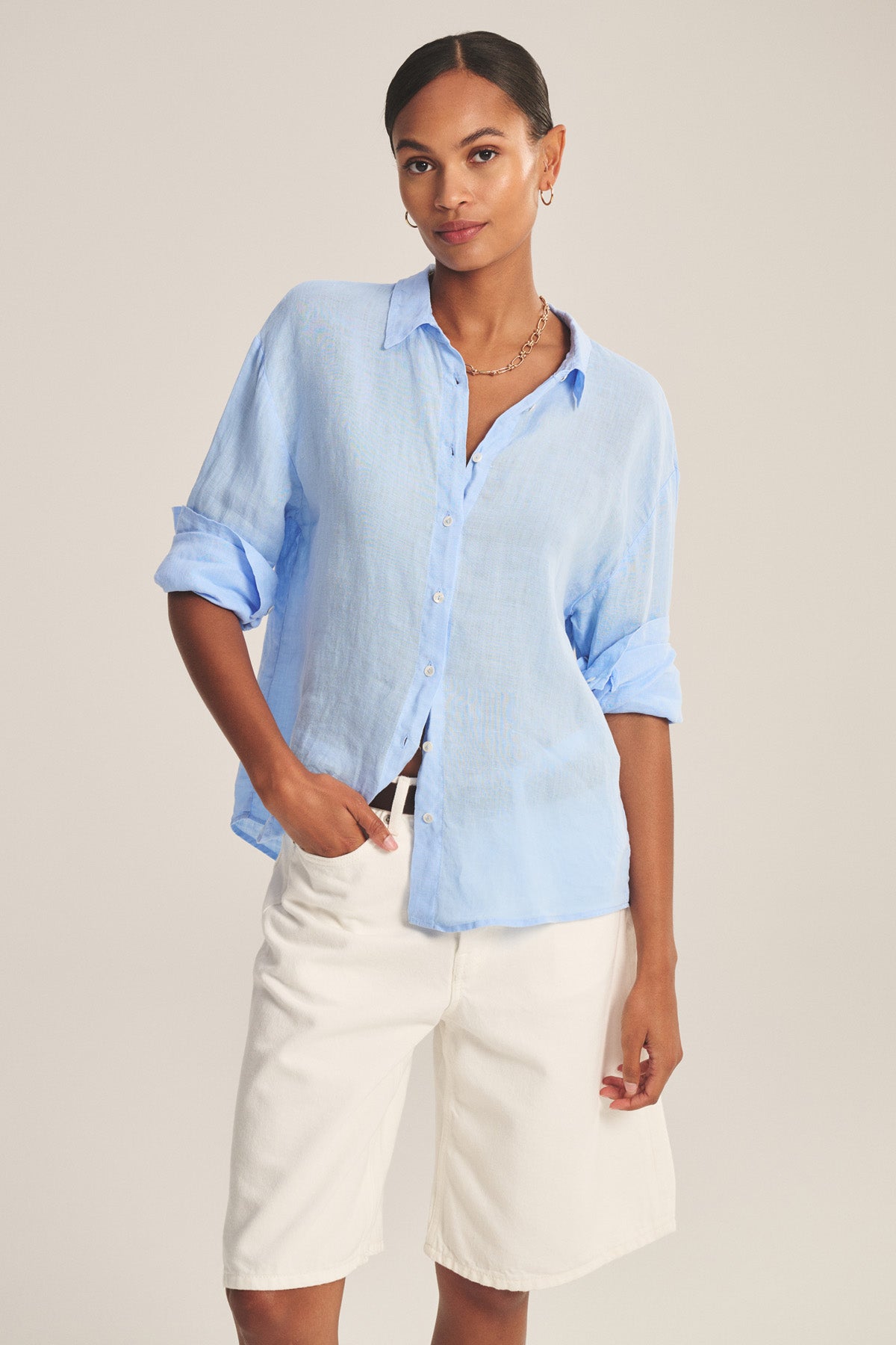 Woman wearing the BRIGHTON LINEN SHIRT by Velvet by Graham & Spencer—a relaxed fit, light blue linen button-up with rolled sleeves—paired with white knee-length shorts, standing against a plain background.