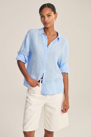 Woman wearing the BRIGHTON LINEN SHIRT by Velvet by Graham & Spencer—a relaxed fit, light blue linen button-up with rolled sleeves—paired with white knee-length shorts, standing against a plain background.