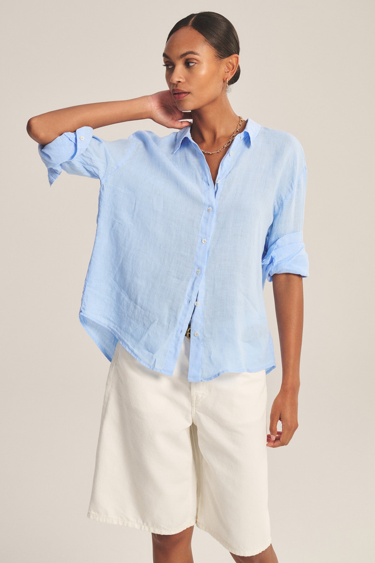 A person wears the BRIGHTON LINEN SHIRT by Velvet by Graham & Spencer, a light blue breathable woven linen button-up with rolled sleeves, paired with white knee-length shorts, standing against a plain background.