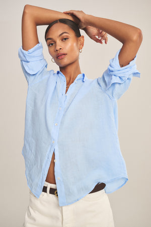 Model wears the BRIGHTON LINEN SHIRT by Velvet by Graham & Spencer, a light blue breathable button-up, paired with white pants, posing with arms raised and crossed above head against a neutral background.
