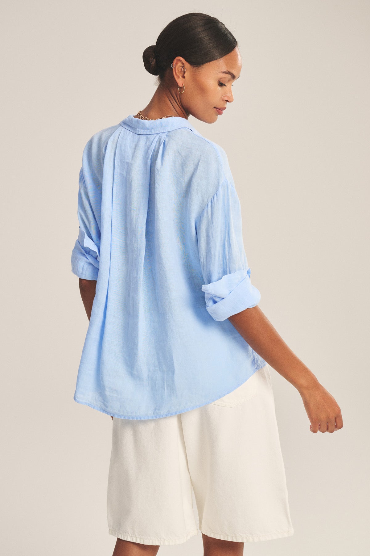 A woman faces away, wearing the Velvet by Graham & Spencer BRIGHTON LINEN SHIRT in light blue with rolled sleeves and a relaxed fit, paired with loose white shorts; her dark hair is tied back in a low bun.