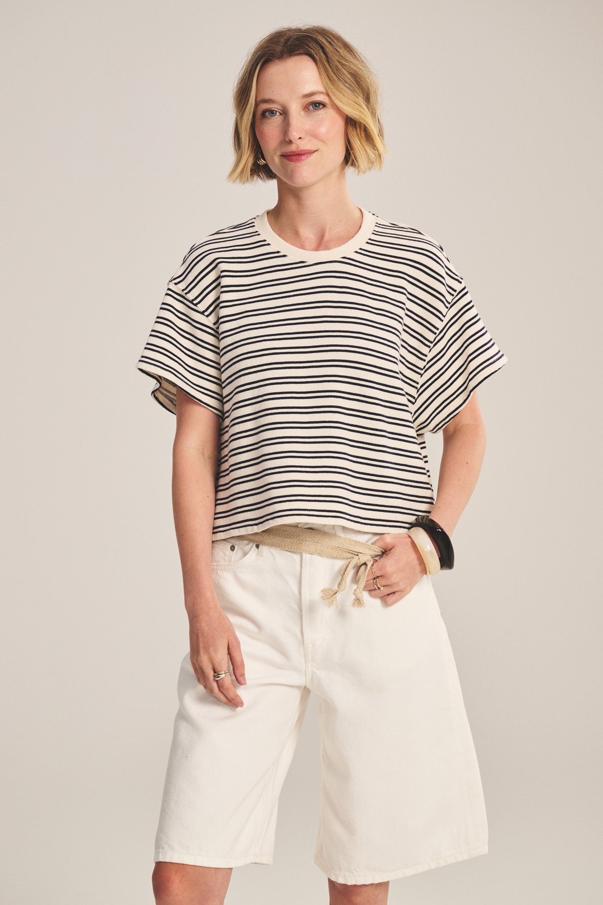 A woman wears the BRISSA TOP by Velvet by Graham & Spencer, a vintage stripe cotton boxy swing pullover, paired with loose white knee-length shorts, one hand in her pocket, exuding relaxed style against a plain background.-40909700726977