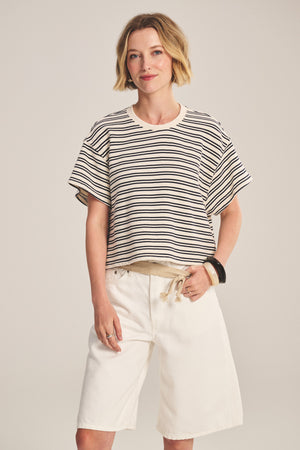 A woman wears the BRISSA TOP by Velvet by Graham & Spencer, a vintage stripe cotton boxy swing pullover, paired with loose white knee-length shorts, one hand in her pocket, exuding relaxed style against a plain background.