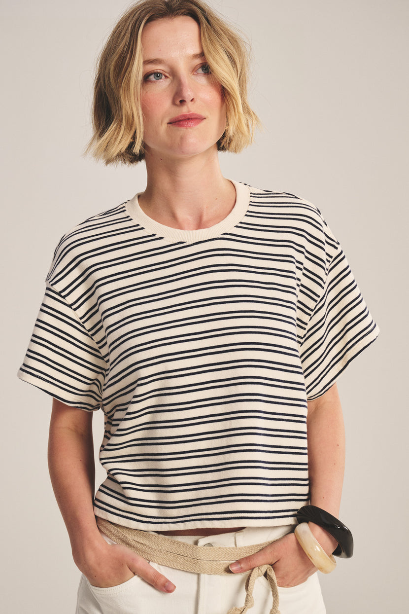 A woman with short blonde hair wears the BRISSA TOP by Velvet by Graham & Spencer— a relaxed fit, vintage stripe cotton t-shirt—paired with white pants and a black bangle, standing against a plain background.