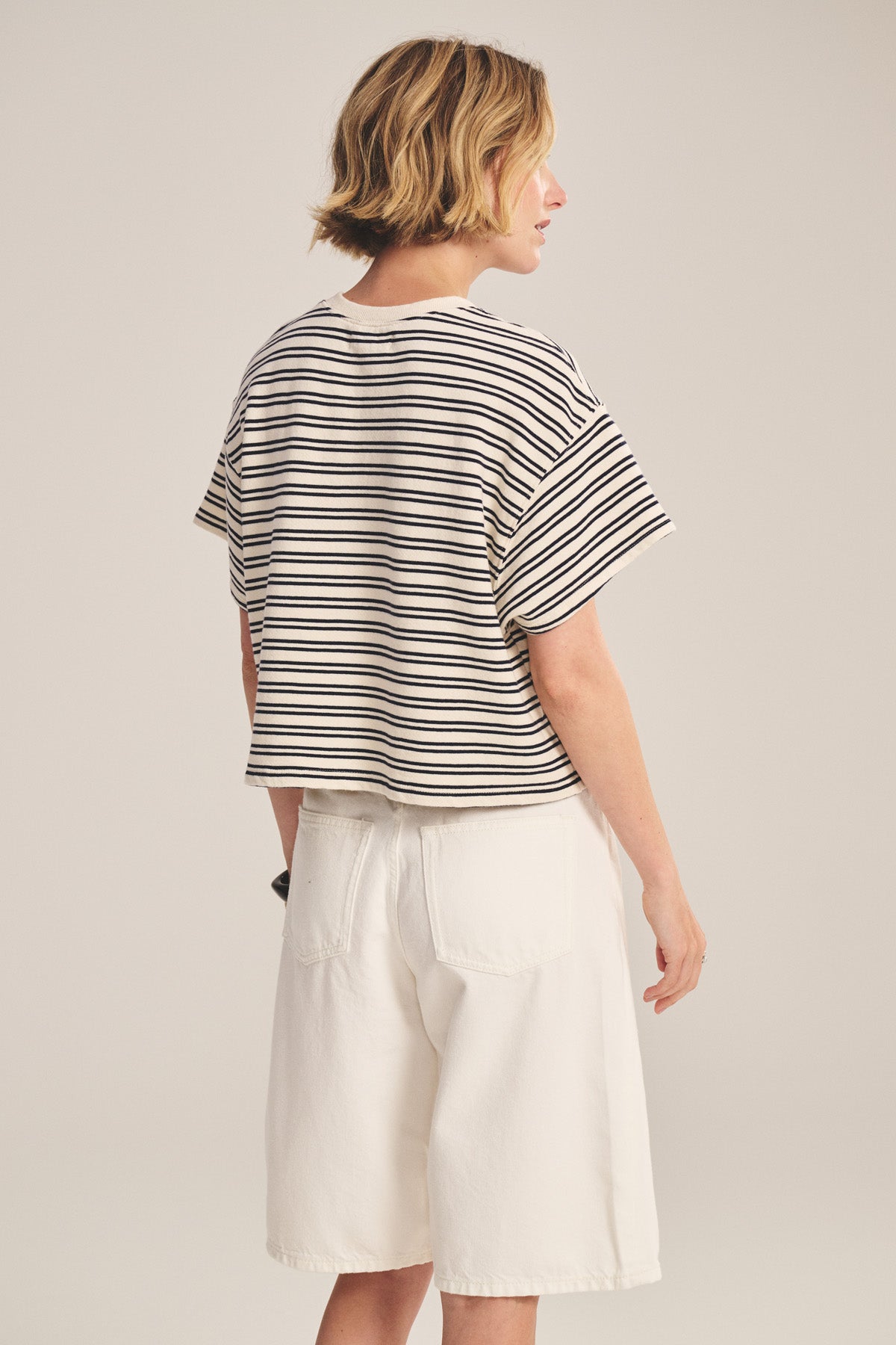 A person stands with their back turned, wearing the BRISSA TOP by Velvet by Graham & Spencer—a vintage stripe cotton short-sleeve in a relaxed fit—paired with loose-fitting white shorts.