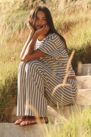A woman with long hair sits on outdoor steps in natural sunlight, wearing the AMELISE SWEATPANT by Velvet by Graham & Spencer—a vintage stripe terry pant with a relaxed straight-leg cut—surrounded by tall grass.