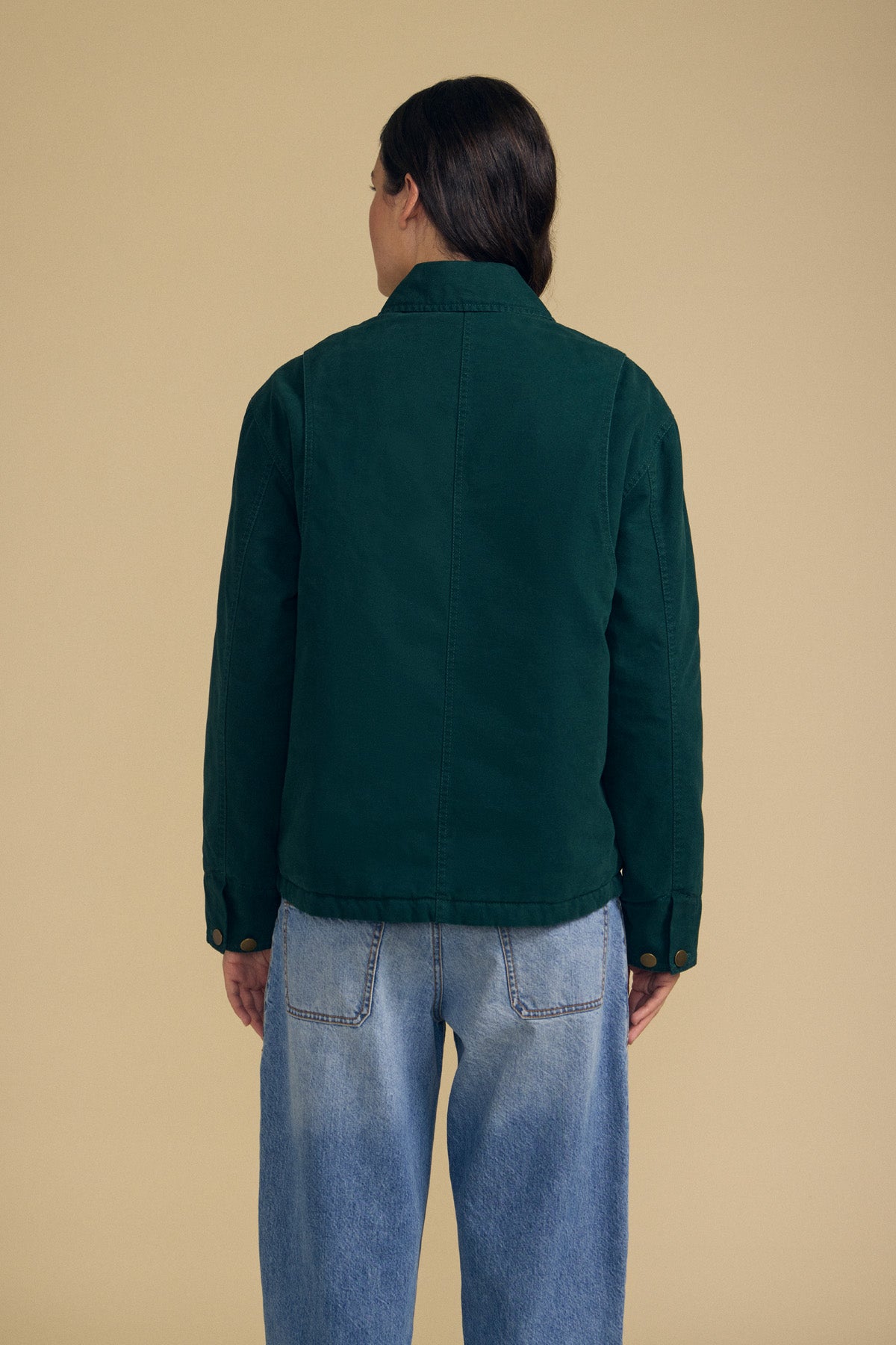 A person stands with their back to the camera, wearing the Velvet by Graham & Spencer DYLAN BARN JACKET in dark green and light blue jeans, set against a plain beige background.