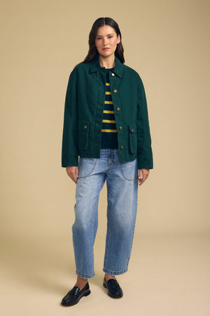 A person stands against a beige background wearing the DYLAN BARN JACKET by Velvet by Graham & Spencer, styled with a striped shirt, light blue cuffed jeans, and black loafers.