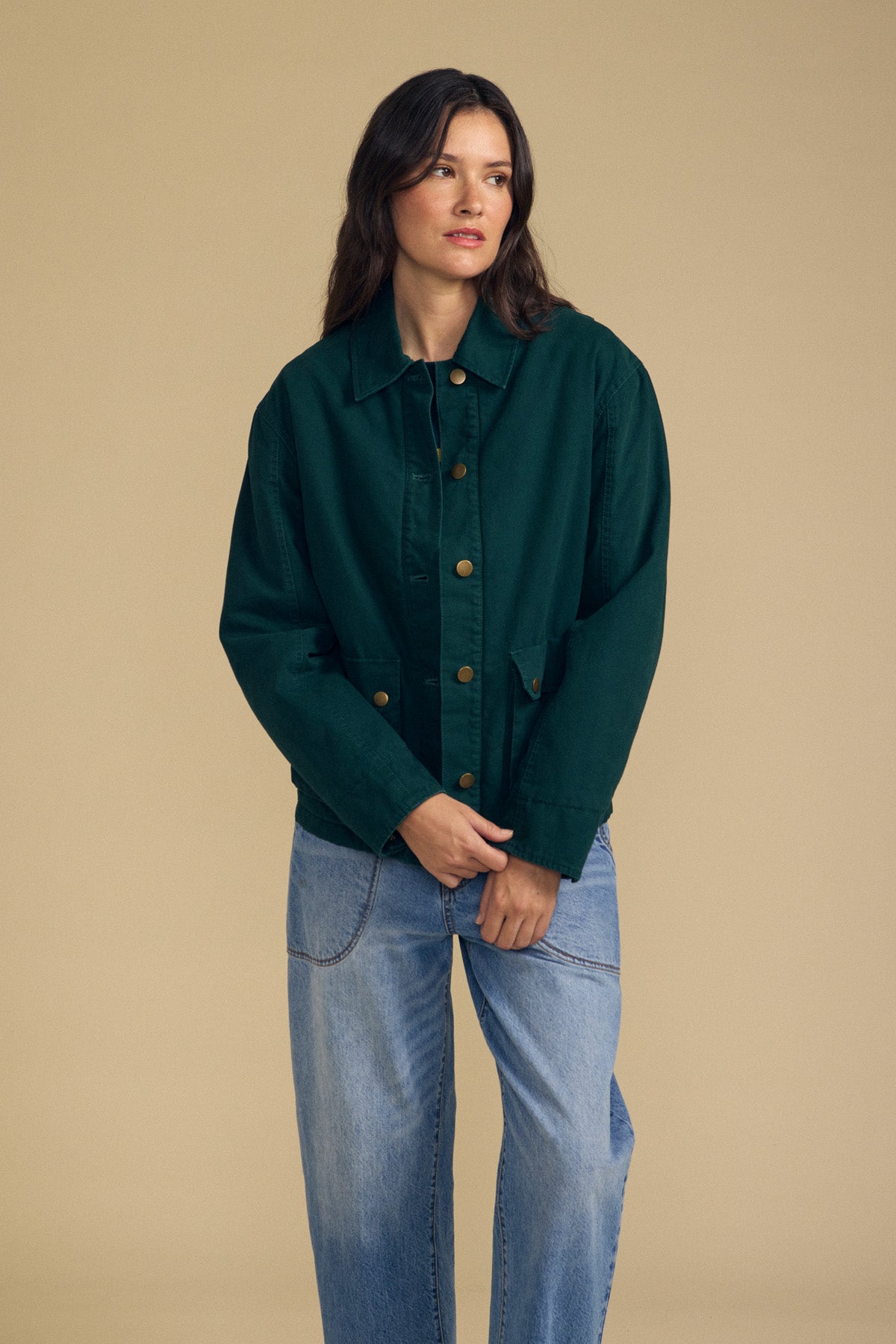 A woman stands against a beige background wearing the Velvet by Graham & Spencer DYLAN BARN JACKET in dark green, paired with light blue jeans and her hands clasped in front of her.