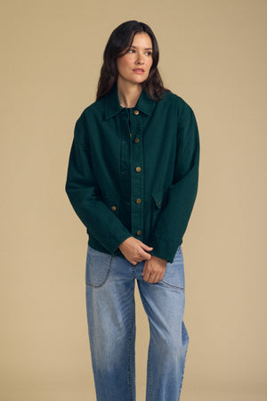 A woman stands against a beige background wearing the Velvet by Graham & Spencer DYLAN BARN JACKET in dark green, paired with light blue jeans and her hands clasped in front of her.