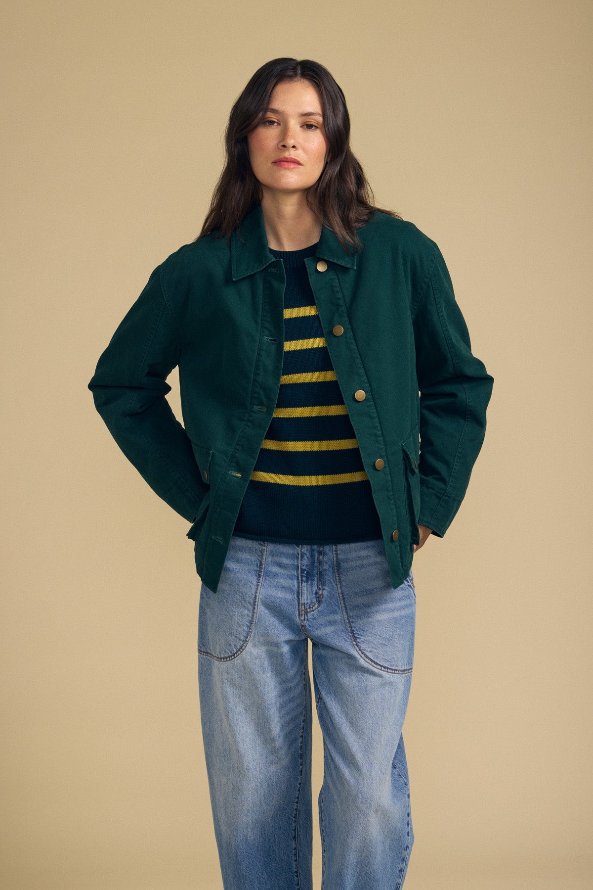 A person wearing the DYLAN BARN JACKET by Velvet by Graham & Spencer over a black and yellow striped sweater with blue jeans stands against a plain beige background.