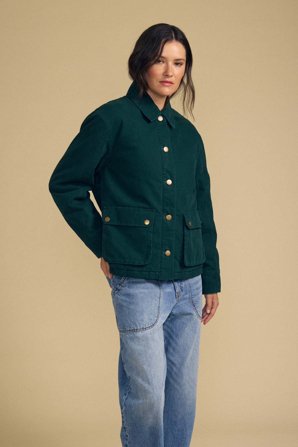 A person wearing the DYLAN BARN JACKET by Velvet by Graham & Spencer—a dark green button-up jacket with front pockets—paired with light blue jeans, stands against a plain beige background.
