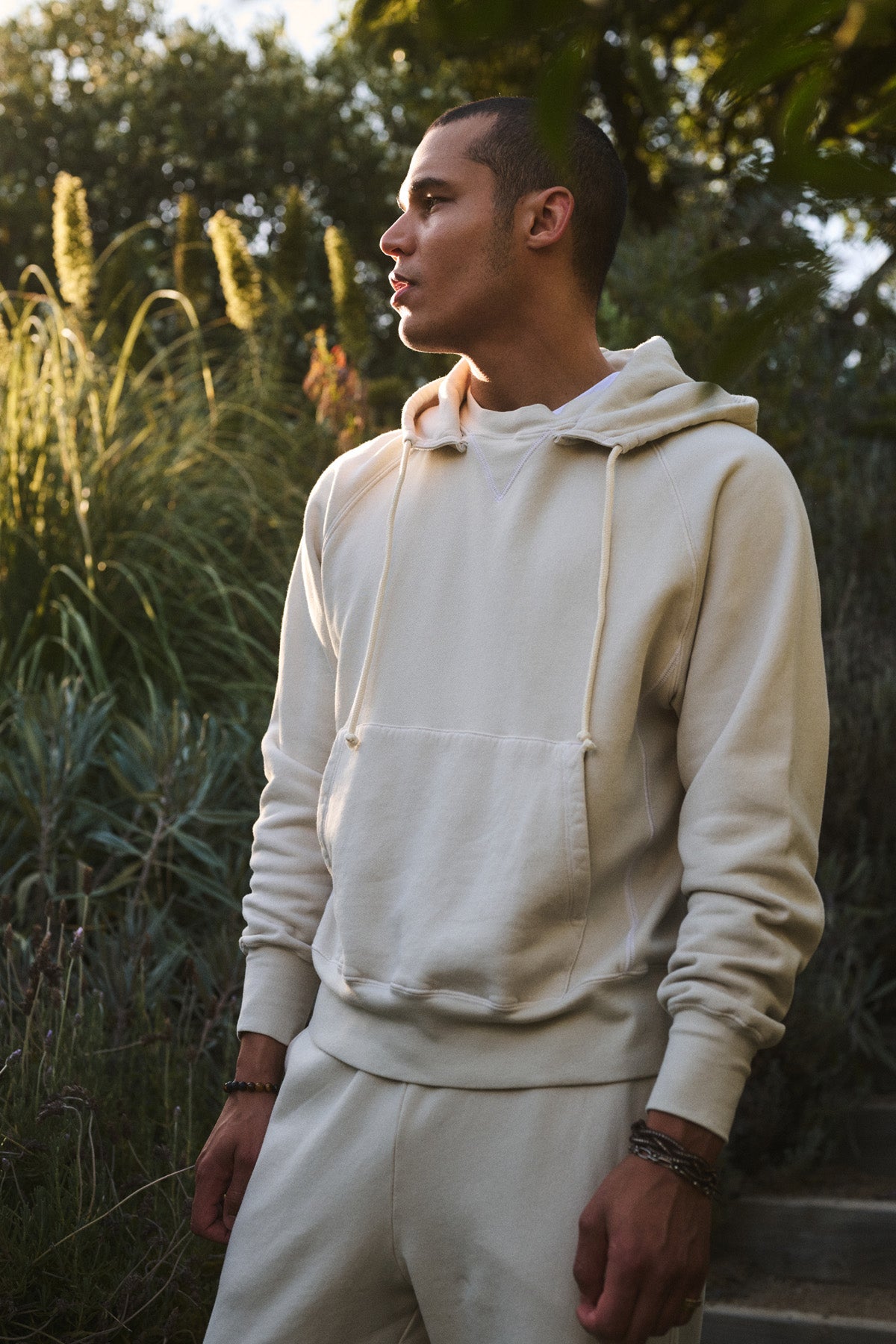 Someone wearing the BRODIE HOODIE by Velvet by Graham & Spencer stands outdoors in natural sunlight, surrounded by greenery. The relaxed fit hoodie features a drawstring hood and is paired with matching joggers.-40850658459841