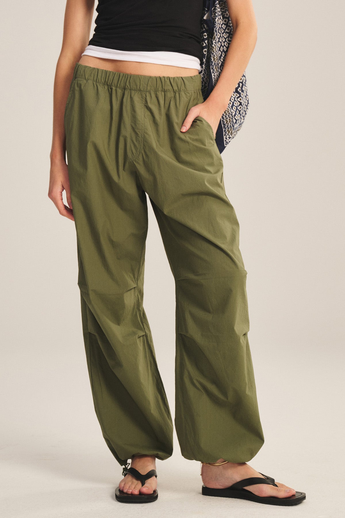 Wearing the BROOKS PANT by Velvet by Graham & Spencer in olive, this person pairs them with a black tank over a white shirt, black sandals, and a patterned bag, standing casually with one hand in their pocket.-41338477019329