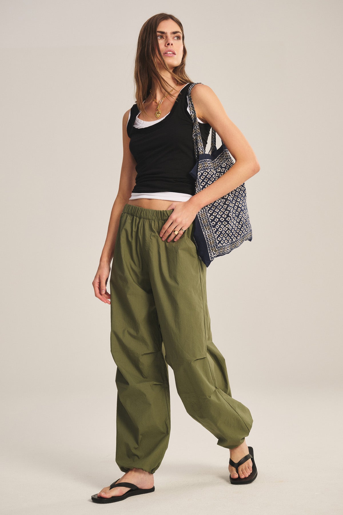 A woman is wearing the BROOKS PANT by Velvet by Graham & Spencer—loose olive green parachute pants with a billowy fit—paired with a black tank top, white shirt, black sandals, and a patterned tote bag over her shoulder.