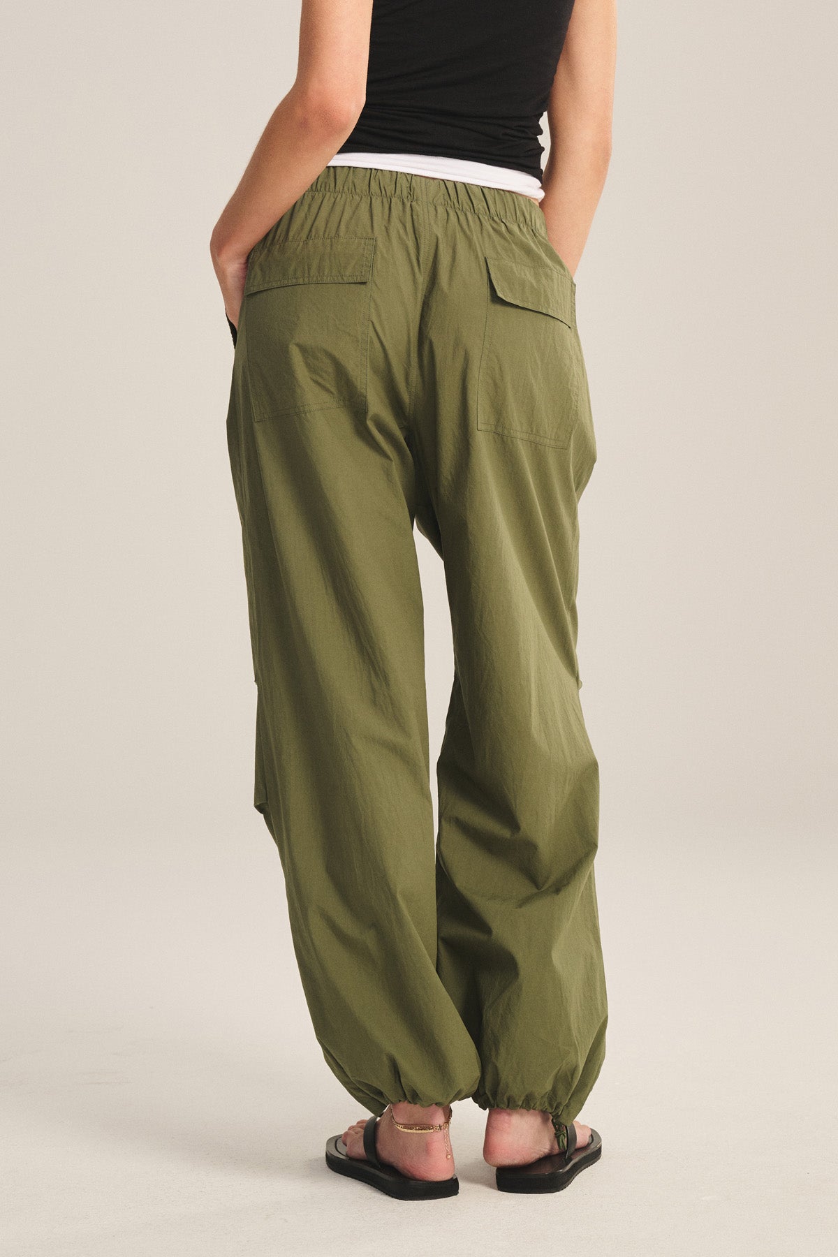 Wearing the BROOKS PANT by Velvet by Graham & Spencer in olive green with a billowy fit, paired with a black top and black sandals, a person stands with hands in back pockets, shown from behind.-41338476921025