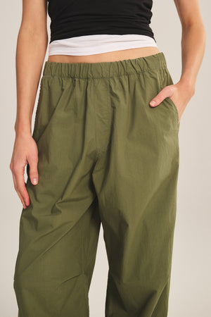 Wearing the BROOKS PANT by Velvet by Graham & Spencer in olive green with a billowy fit and elastic waistband, paired with a black top, and standing with one hand in their pocket.