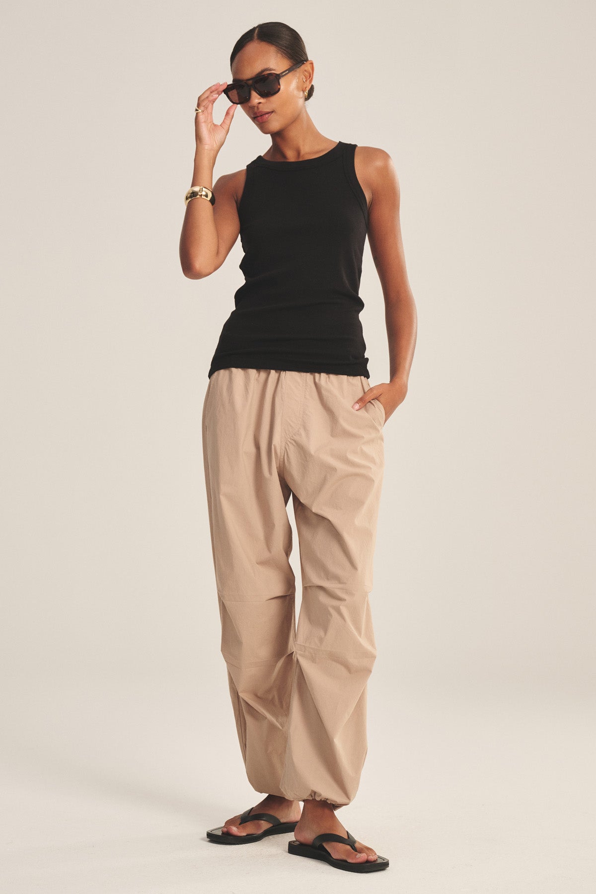 Wearing a black sleeveless top, Velvet by Graham & Spencer's BROOKS PANT in cotton poplin with parachute silhouette and cinched corded cuffs, black sandals, and sunglasses, a person stands with one hand in their pocket.