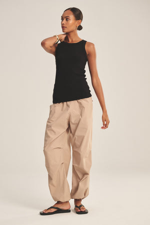 A person stands against a plain background wearing a black sleeveless top, Velvet by Graham & Spencer’s BROOKS PANT with a parachute silhouette, black sandals, and a bracelet, one hand resting on their neck.