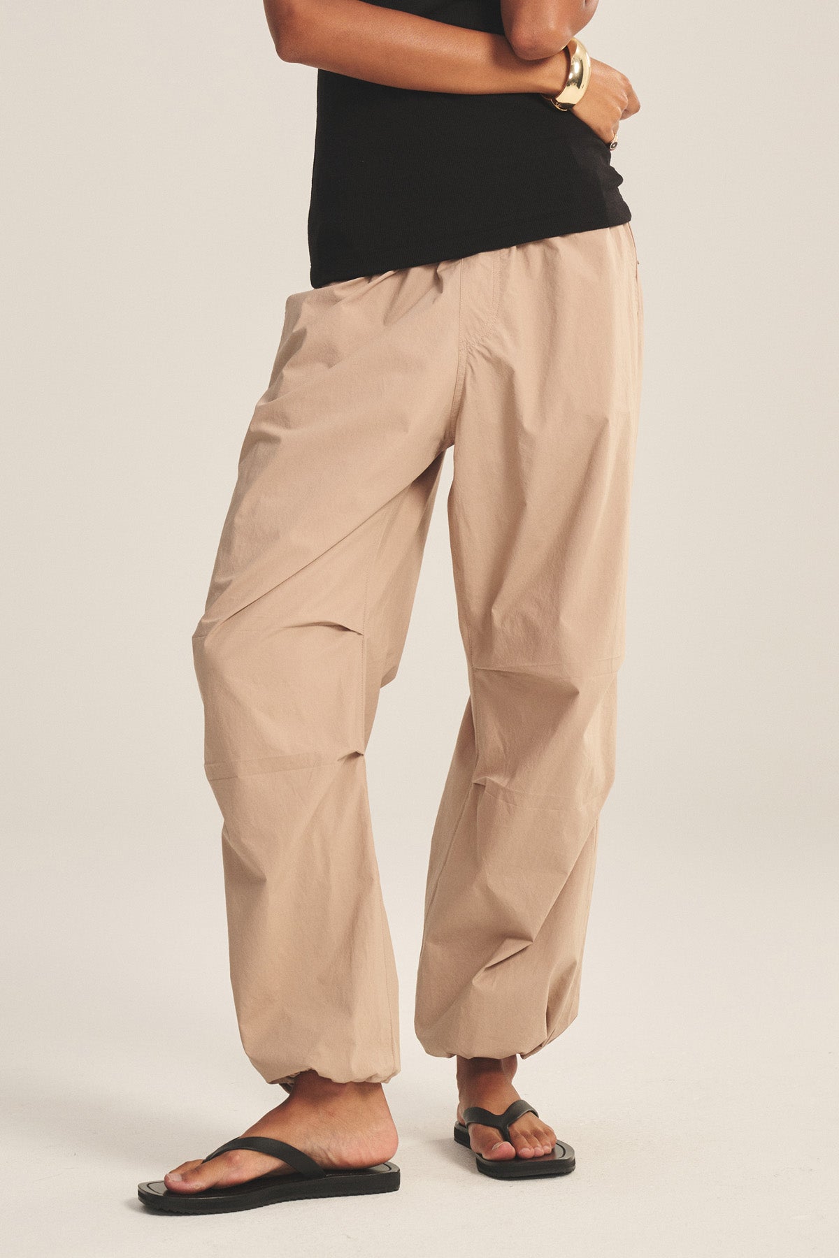 A person wears the BROOKS PANT by Velvet by Graham & Spencer—beige cotton poplin joggers with cinched corded cuffs—paired with a black top, bracelet, and black sandals, standing against a plain light background.-41081163219137