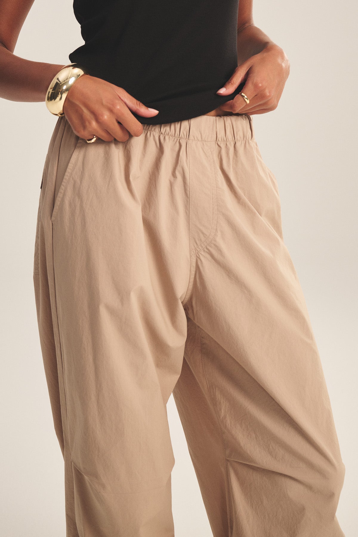 A person wearing the BROOKS PANT by Velvet by Graham & Spencer—beige cotton poplin with cinched corded cuffs—paired with a black sleeveless top and gold bracelet, hands resting at the waistband.-41081163186369