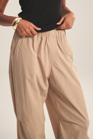 A person wearing the BROOKS PANT by Velvet by Graham & Spencer—beige cotton poplin with cinched corded cuffs—paired with a black sleeveless top and gold bracelet, hands resting at the waistband.