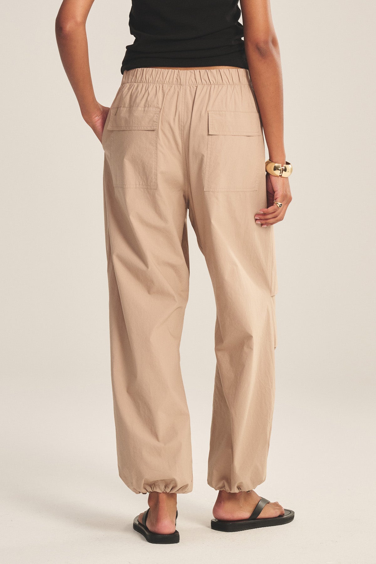 A person viewed from the back wearing Velvet by Graham & Spencer’s BROOKS PANT in beige cotton poplin with corded cuffs and patch back pockets, paired with a black top, black sandals, and a gold bracelet.
