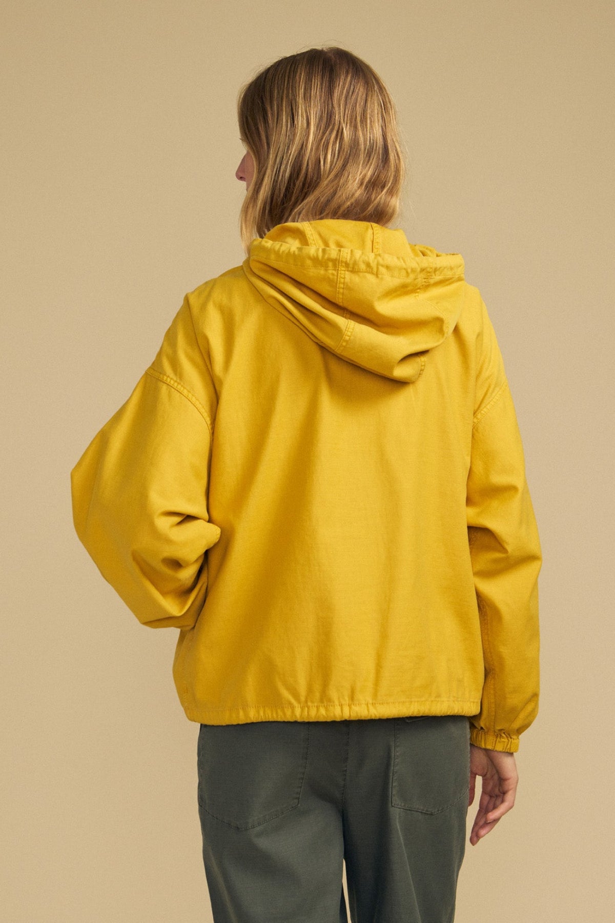 Wearing the BOBBIE HOODIE by Velvet by Graham & Spencer—an oversized, brushed cotton yellow hooded jacket—paired with green pants, a person stands facing away against a plain beige background.-41970970198209