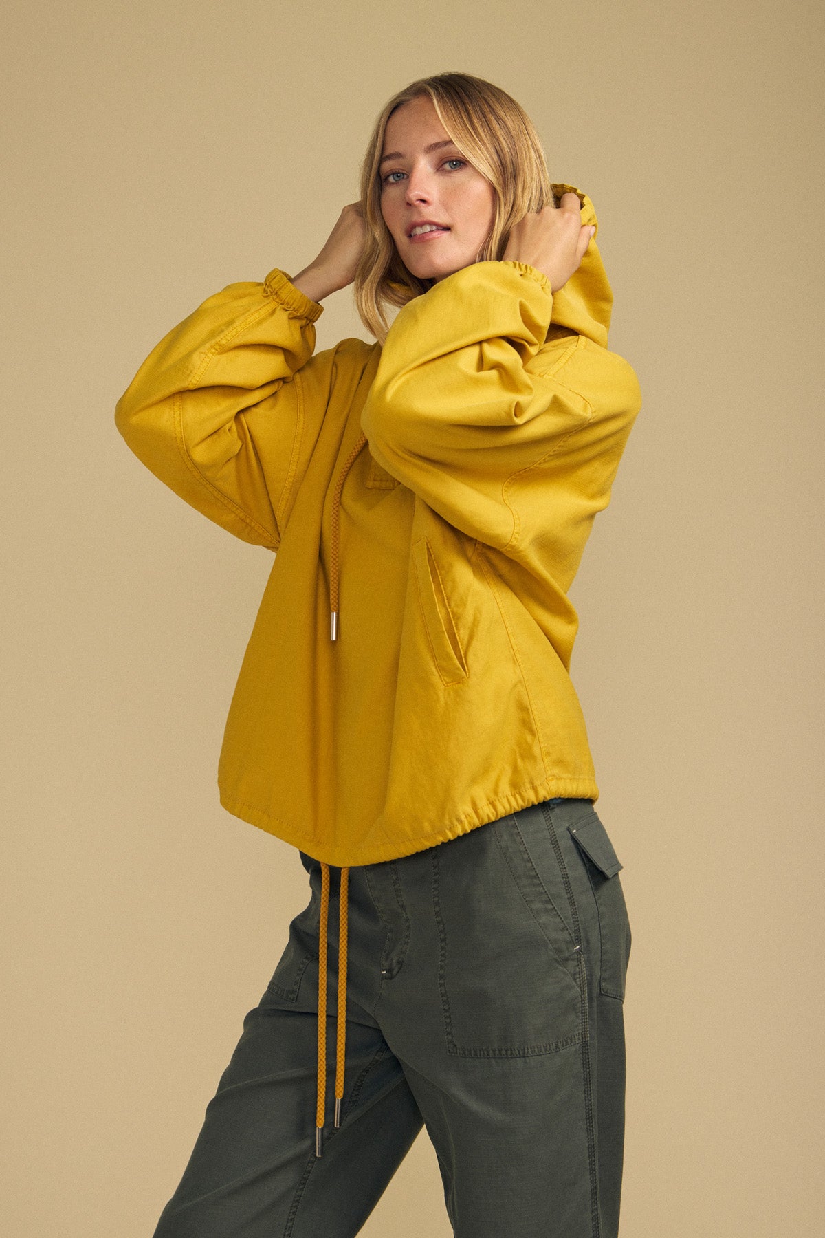 A person wears the Velvet by Graham & Spencer BOBBIE HOODIE in yellow, featuring an oversized silhouette, paired with olive green pants, standing against a plain beige background and looking at the camera.