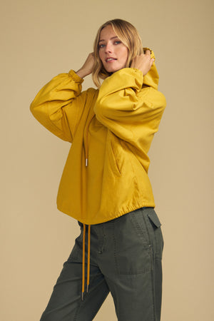 A person wears the Velvet by Graham & Spencer BOBBIE HOODIE in yellow, featuring an oversized silhouette, paired with olive green pants, standing against a plain beige background and looking at the camera.