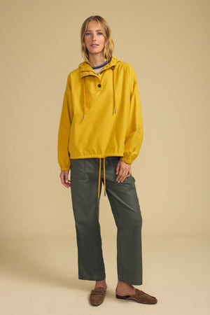 Wearing the BOBBIE HOODIE by Velvet by Graham & Spencer, a person stands against a plain beige background. The yellow hooded jacket features an oversized silhouette and is paired with green pants and brown slip-on shoes.