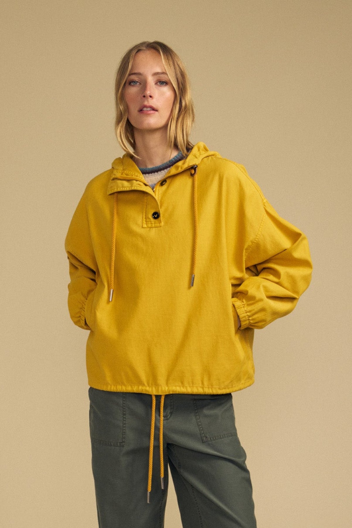 A woman wears the BOBBIE HOODIE by Velvet by Graham & Spencer—a loose-fitting yellow hooded jacket with drawstrings in an oversized silhouette—paired with green cargo pants, standing against a beige background.