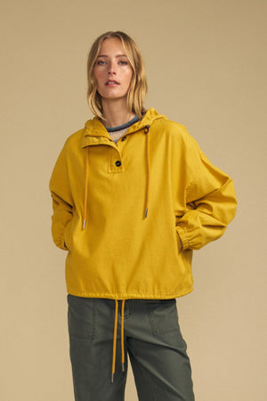 A woman wears the BOBBIE HOODIE by Velvet by Graham & Spencer—a loose-fitting yellow hooded jacket with drawstrings in an oversized silhouette—paired with green cargo pants, standing against a beige background.