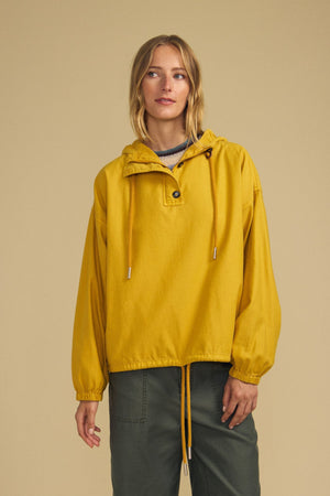 A person stands against a beige background wearing the Velvet by Graham & Spencer BOBBIE HOODIE, a loose-fitting yellow pullover with a drawstring and olive green pants, showcasing an oversized silhouette in soft brushed cotton.