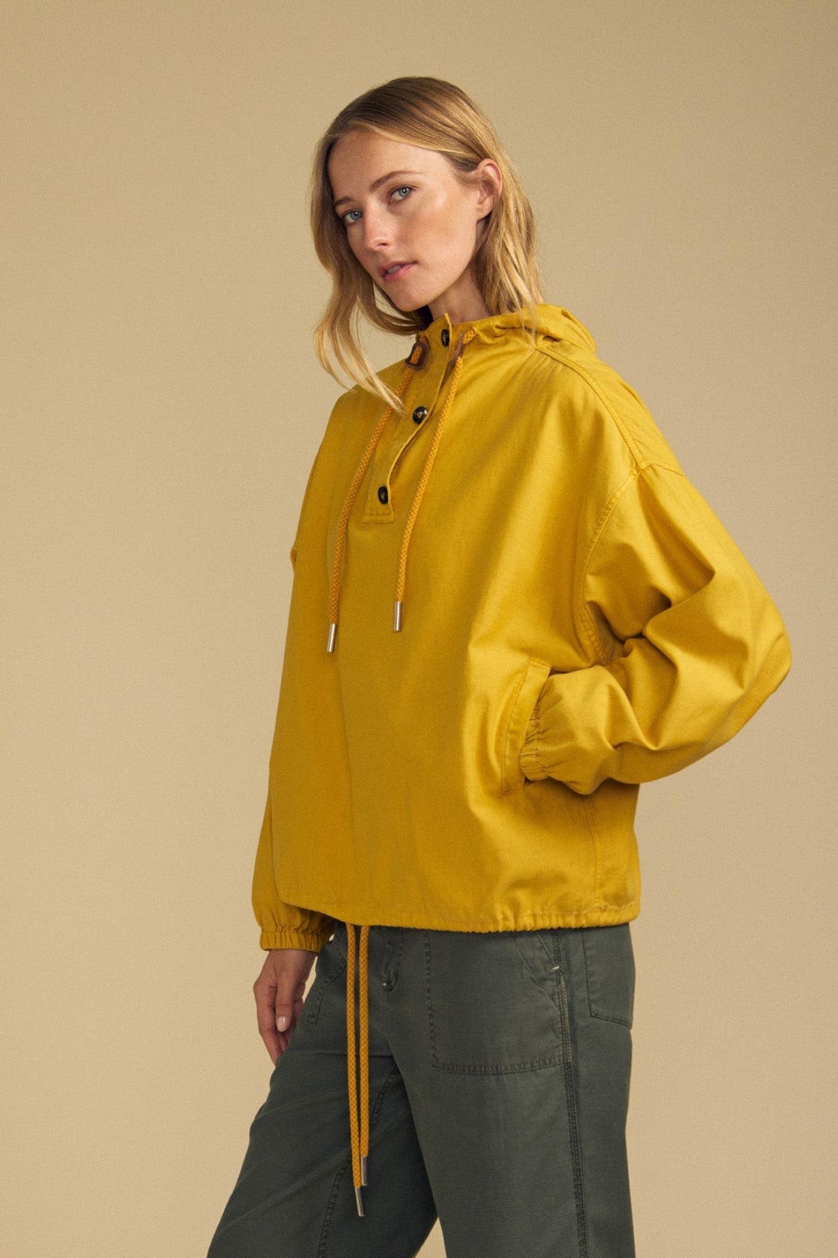 A person wears the BOBBIE HOODIE by Velvet by Graham & Spencer—a yellow, oversized hooded jacket with drawstrings, crafted from soft brushed cotton—paired with green pants and standing against a beige background.