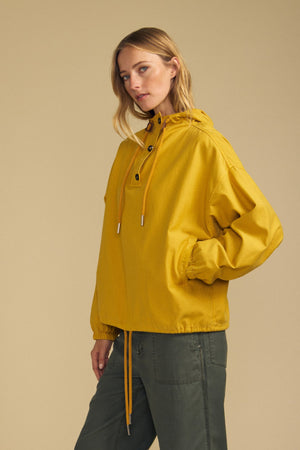 A person wears the BOBBIE HOODIE by Velvet by Graham & Spencer—a yellow, oversized hooded jacket with drawstrings, crafted from soft brushed cotton—paired with green pants and standing against a beige background.