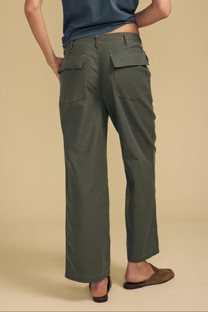 A person stands with their back to the camera, wearing the JACLYN PANT in olive green brushed cotton sateen by Velvet by Graham & Spencer, paired with a blue t-shirt and brown slip-on shoes, set against a plain beige background.