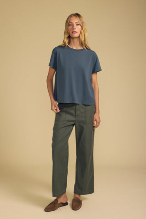 A woman stands against a beige background wearing a blue short-sleeve t-shirt, the JACLYN PANT in olive brushed cotton sateen by Velvet by Graham & Spencer, and brown slip-on shoes.