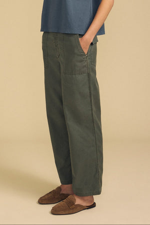A person wearing the JACLYN PANT by Velvet by Graham & Spencer, styled in a loose-fitting green wide-leg silhouette, along with a blue top and brown loafers, stands with hands in pockets against a beige background.
