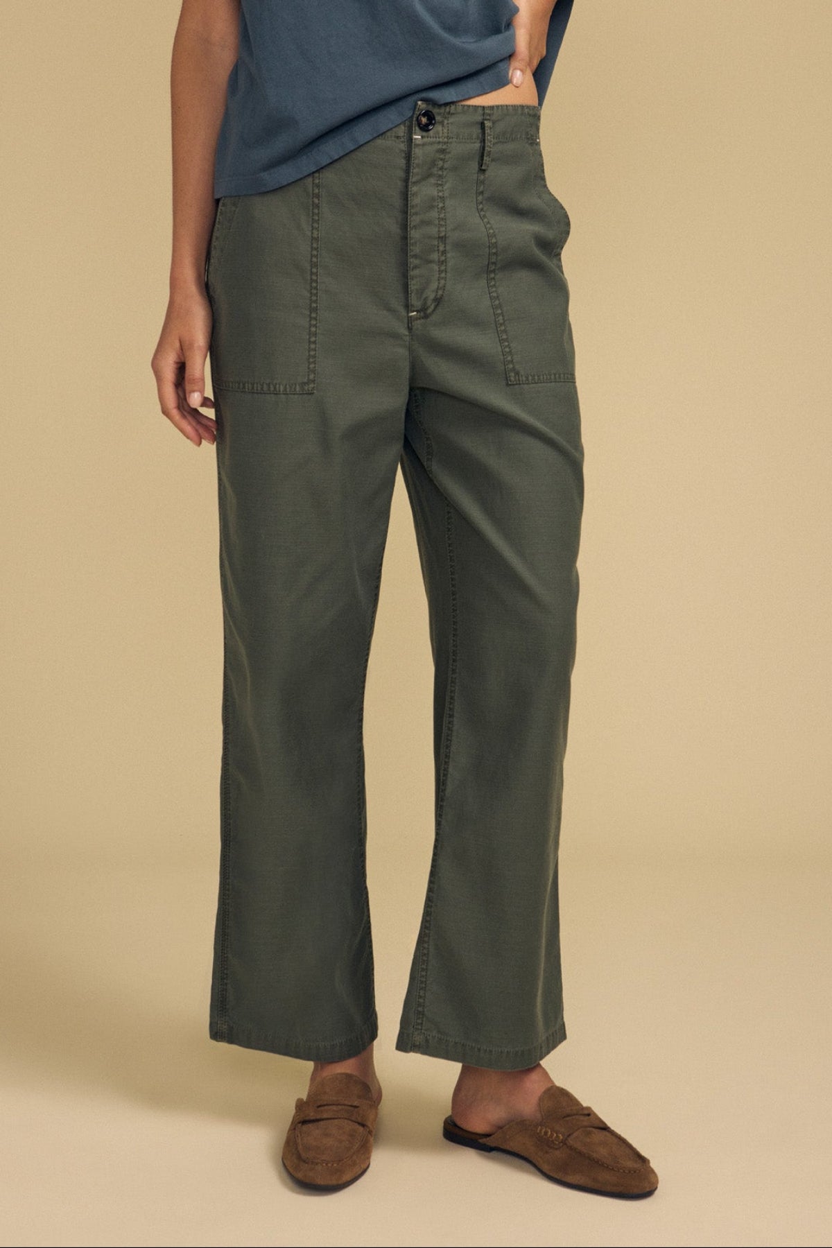 A person stands with one hand on their hip, wearing a blue t-shirt, brown slip-on shoes, and the JACLYN PANT by Velvet by Graham & Spencer—an olive green wide-leg pant in brushed cotton sateen with front pockets—against a beige background.