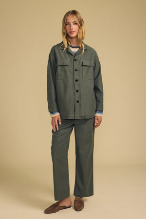 A woman stands facing forward against a beige background, wearing the JACLYN PANT by Velvet by Graham & Spencer—olive green brushed cotton sateen wide-leg pants—paired with a matching button-up jacket and brown loafers.