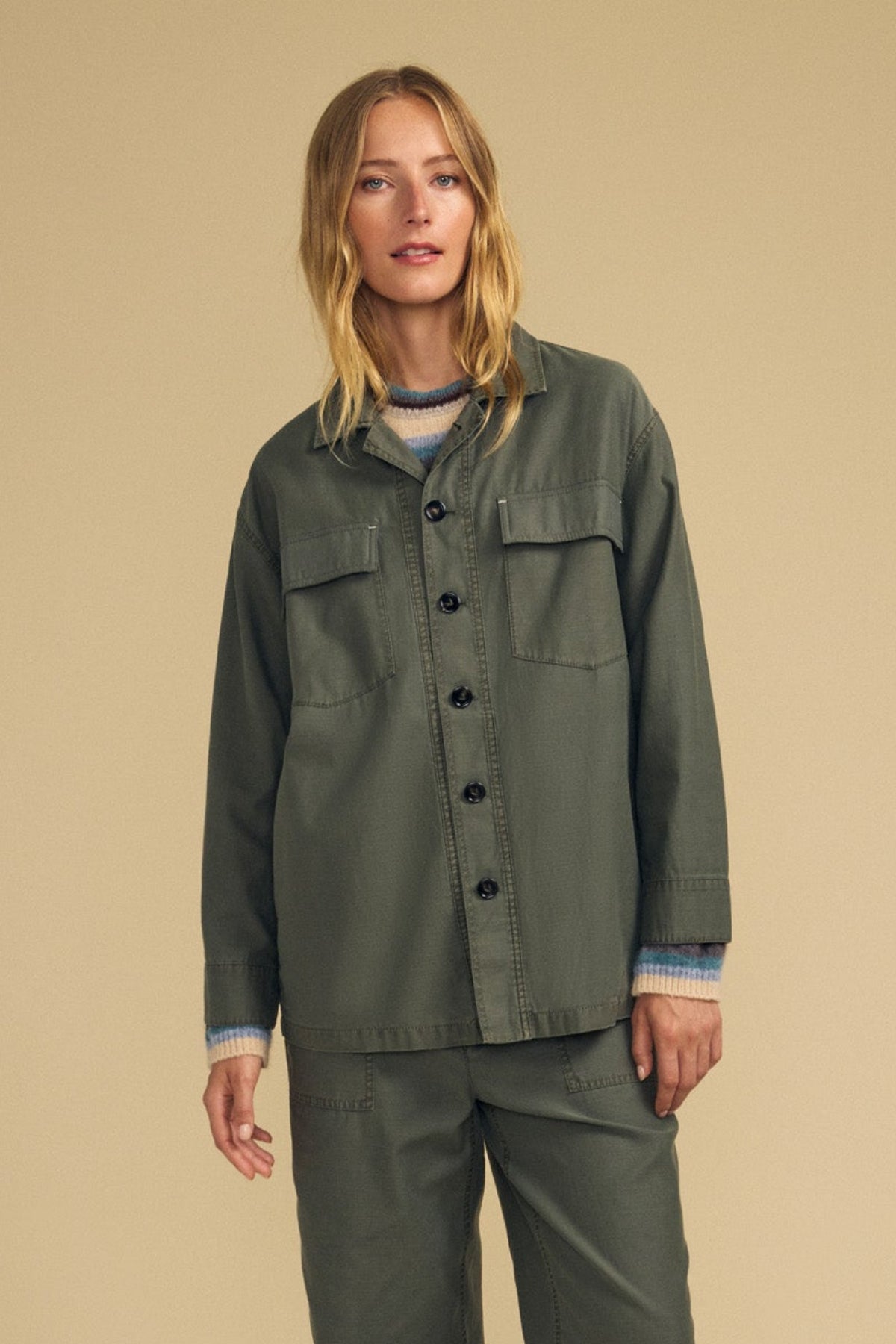 A person stands against a beige background wearing Velvet by Graham & Spencer’s olive green KAY SHIRT JACKET paired with matching pants and a striped sweater underneath.