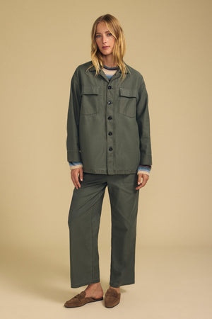 A woman stands facing forward in a beige studio, wearing the KAY SHIRT JACKET by Velvet by Graham & Spencer in olive green brushed cotton, paired with matching wide-leg pants, layered over a blue and white sweater, and finished with brown slip-on shoes.