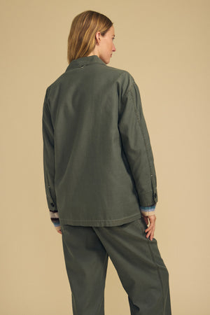 A person with light brown hair stands facing away, wearing the KAY SHIRT JACKET by Velvet by Graham & Spencer in olive green brushed cotton, paired with matching pants, against a plain beige background.