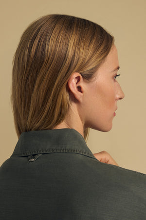 A woman with straight blonde hair, seen from behind and in profile, wears the KAY SHIRT JACKET by Velvet by Graham & Spencer—a dark green brushed cotton button-up—set against a beige background.