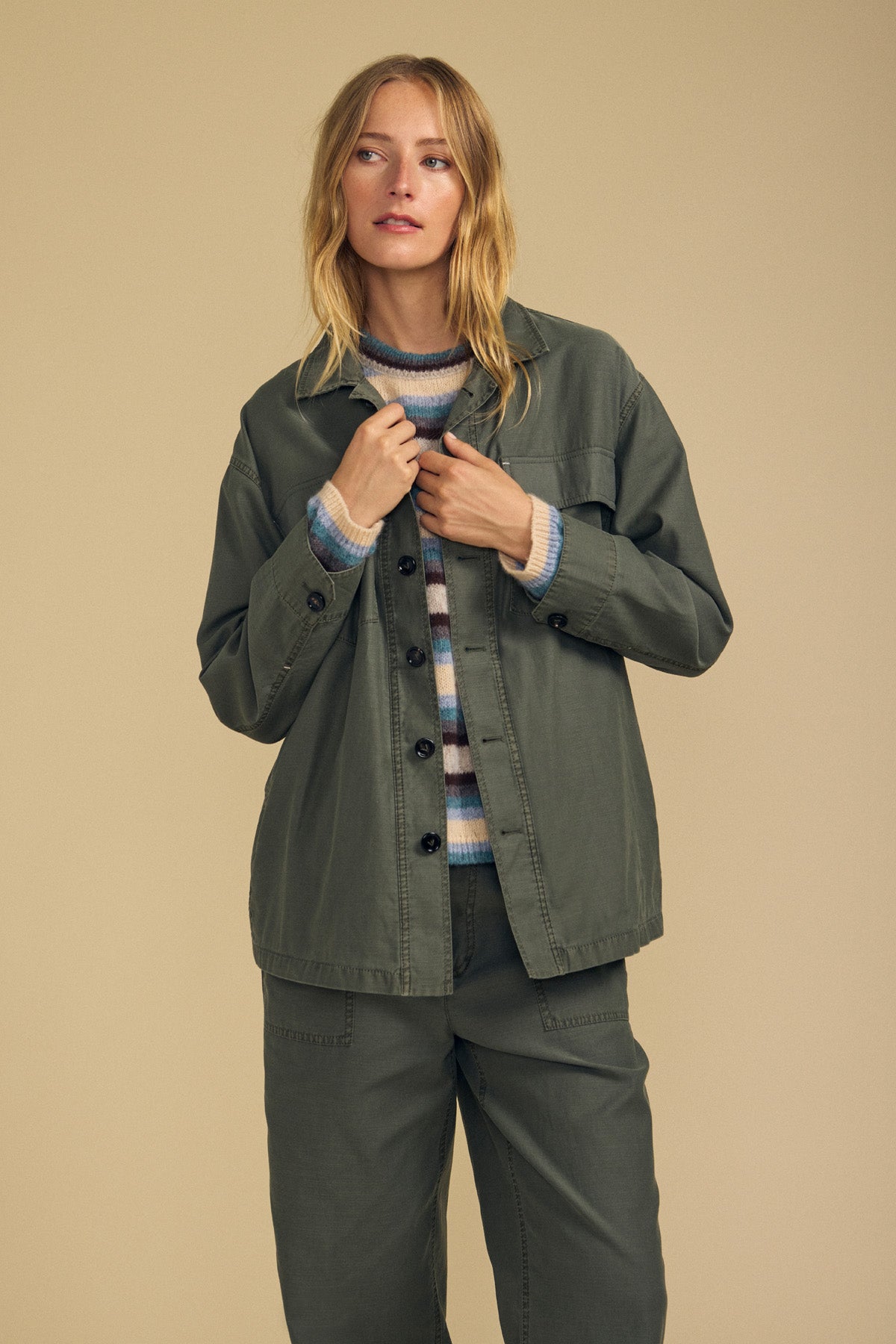 A person stands against a beige background, wearing the KAY SHIRT JACKET by Velvet by Graham & Spencer in olive green paired with matching pants over a striped sweater, looking slightly to the side. This lightweight jacket is crafted from brushed cotton, offering both comfort and style to the outfit.