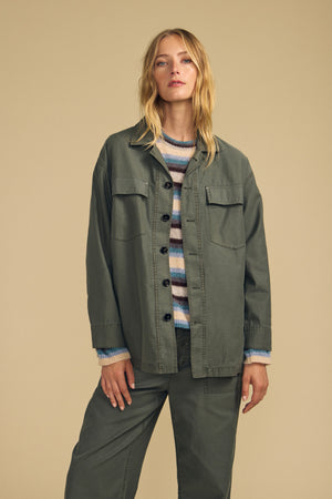 A woman stands against a beige background wearing the KAY SHIRT JACKET in olive green brushed cotton by Velvet by Graham & Spencer, styled over a striped sweater and paired with matching pants.