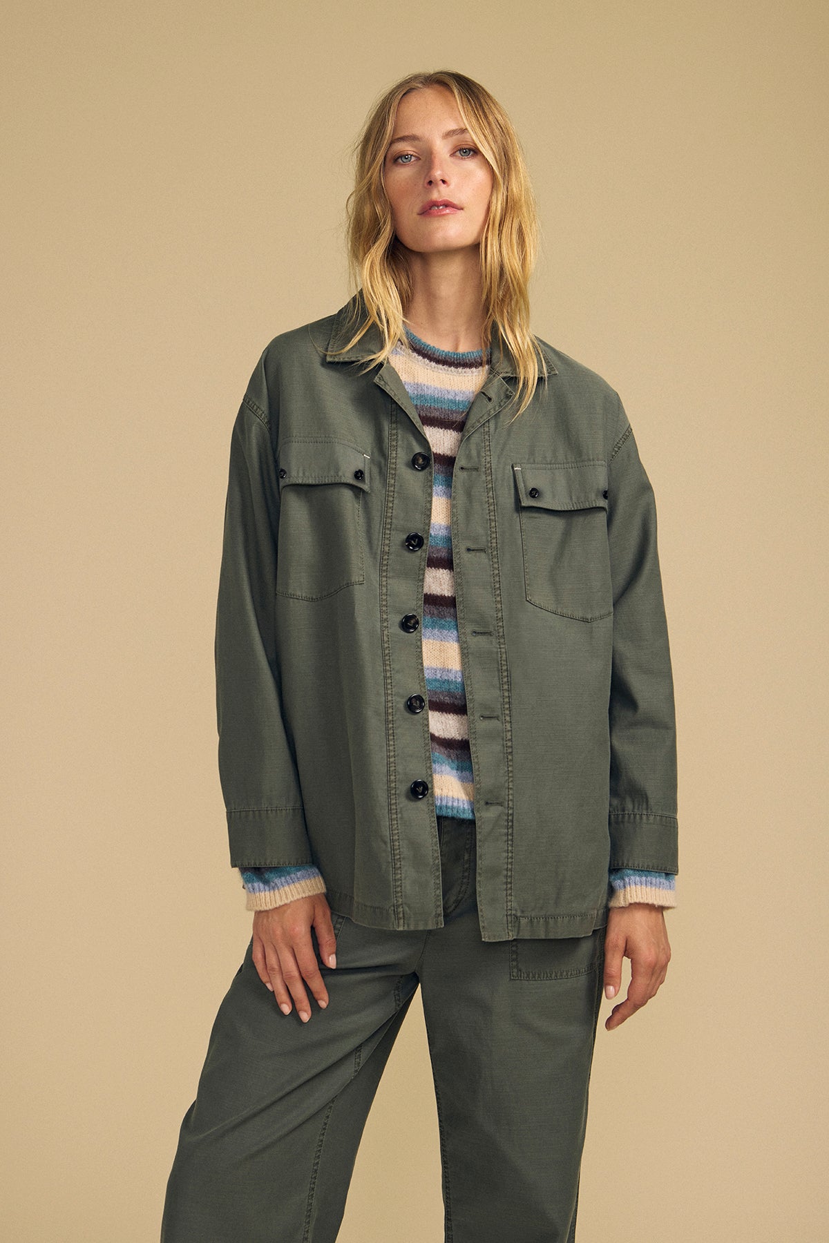 A person wears the KAY SHIRT JACKET by Velvet by Graham & Spencer, styled over a striped sweater and green pants, standing against a plain beige background.