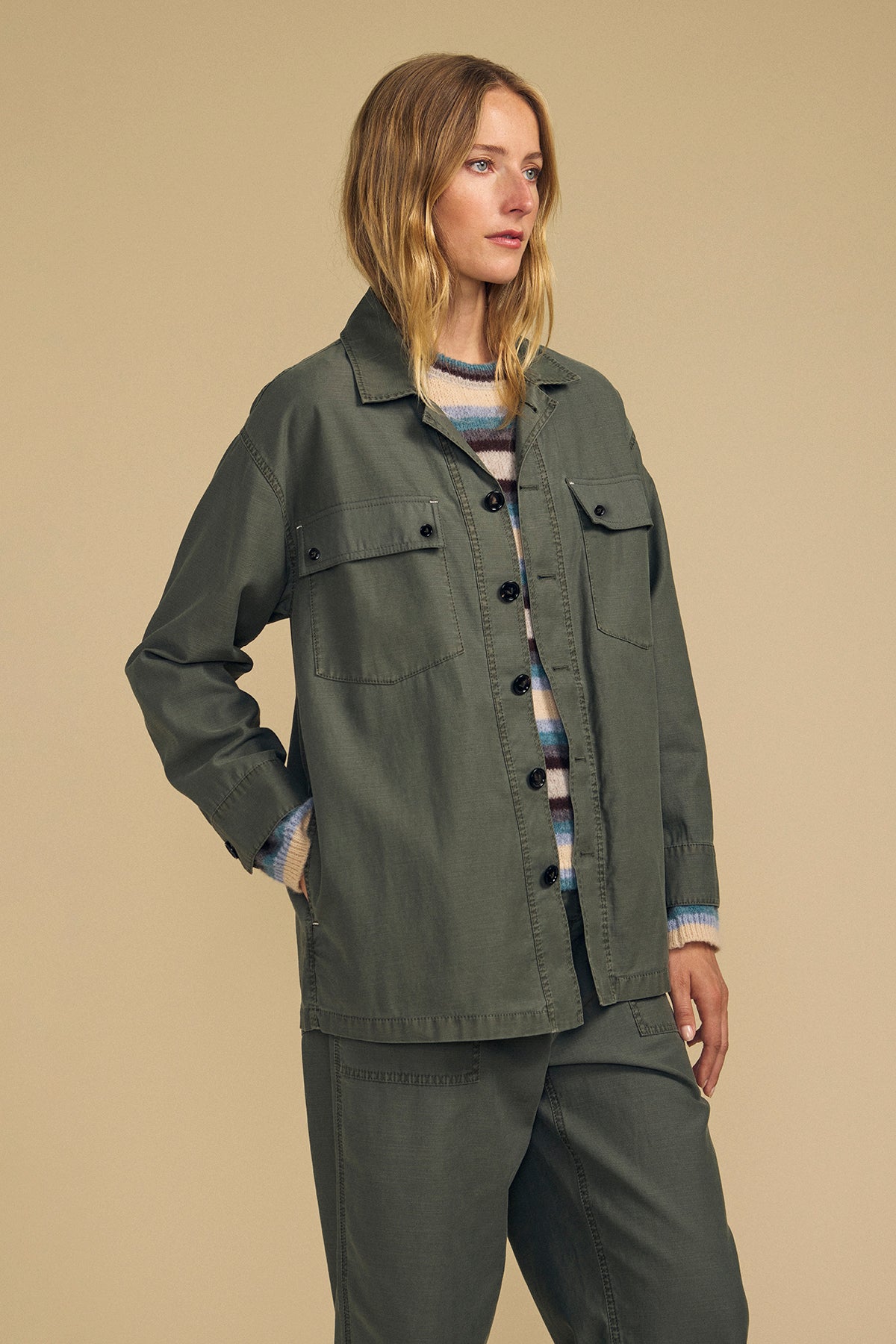 A woman wears the KAY SHIRT JACKET by Velvet by Graham & Spencer with matching pants and a striped sweater, posed against a plain beige background.-46257705058497