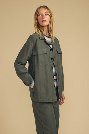 A person stands against a beige background wearing the KAY SHIRT JACKET in olive green brushed cotton by Velvet by Graham & Spencer, paired with matching pants over a striped sweater.